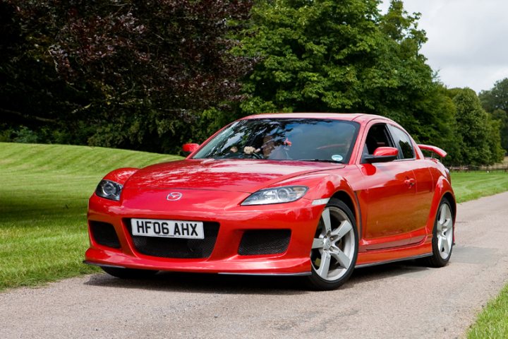 Pistonheads Manor Motors - The image showcases a vibrant red Mazda RX-8 sports car parked on a gravel road, with a mix of shade and sunlight hitting its glossy side. The car is well-maintained and features chrome accents and a distinctive rear design that includes a wing and a white stripe that stands out against the car's red paint. The car's license plate reads "HFO AHX", indicating it's registered in the United Kingdom. In the background, there's a serene landscaped with grass and trees that complement the lively colors of the car.