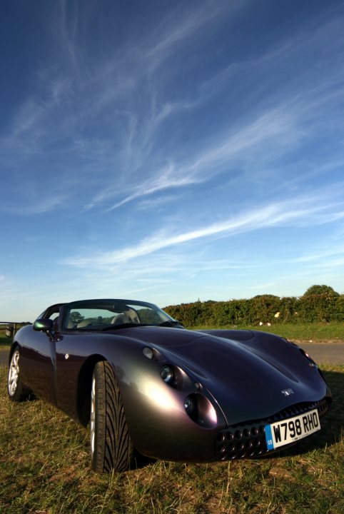 Pistonheads - The image shows a newer model sports car parked on a green grass field. The car is a sleek, dark metal color with a low and wide stance, typical of sports cars. It features a large front grille and air intake vents, along with distinctive styled headlights and taillights, indicative of a modern, aerodynamic design. The sky above is a clear blue with thin, wispy clouds, suggesting a sunny day, which casts a bright light on the car. The surrounding field is spacious and the car is alone in the frame, providing a clean and uncluttered focus on the vehicle.