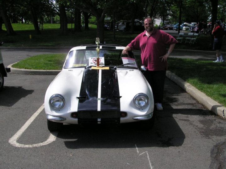 Early TVR Pictures - Page 65 - Classics - PistonHeads - The image shows a man standing next to a vintage race car in a parking lot. The car is predominantly white, with a black stripe along the side. It features a large hood scoop and a prominent front spoiler. The man is wearing a red shirt and glasses. In the front window of the car is a trophy where the man has placed a plaque with an image of the car. In the background, there are other cars parked further away, and the setting appears to be a park or similar outdoor public space.