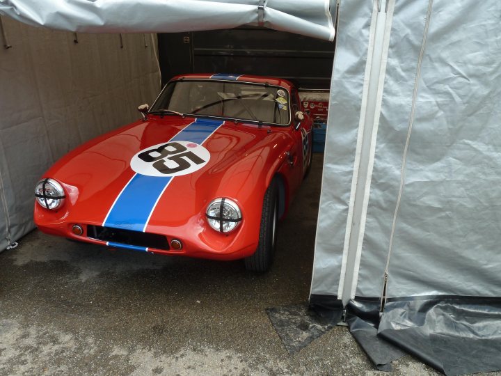 Spa 6 hours, 2015 - Page 1 - Classics - PistonHeads - The image features a classic red sports car with a blue stripe on its side. The car is parked inside a garage with its driver's side door open. The garage has a sturdy metal door and is partially covered by a white tarp. The car is unoccupied and appears to be in good condition, suggesting it is well cared for.