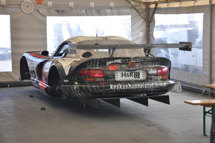 Jim Pistonheads Clark Hockenheimring - The image shows a racecar in a garage or large indoor space. The car is predominantly silver and black, with a large rear wing and European license plates. Writing on the side of the car includes "H&M" and "keshusjune". There's a person partially visible behind the car, and a clock mounted on the wall in the background. The room has windows, and there's another car in the background, suggesting it might be a shared garage or workspace. The overall setting, including the state of dust and wear on the car, implies that the vehicle may be inactive or undergoing maintenance.