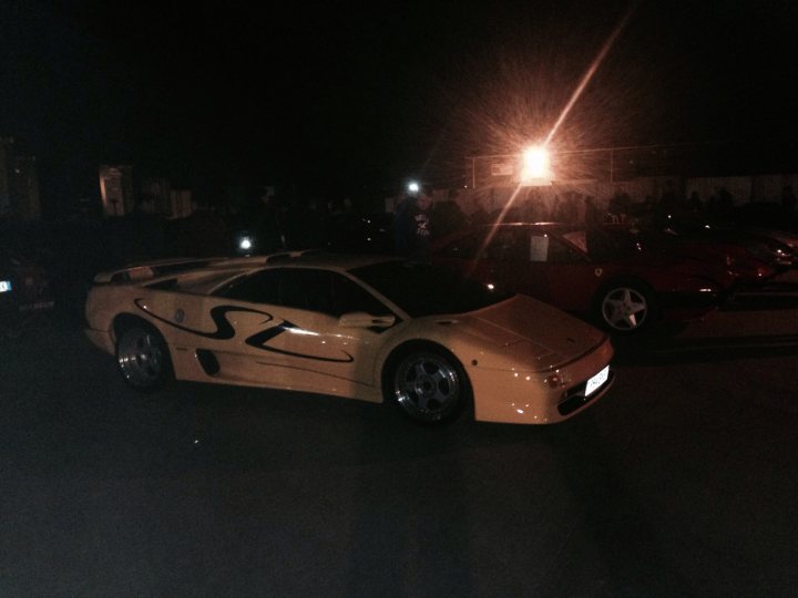 The GT Cafe Gallery - post your meeting photos here - Page 1 - North West - PistonHeads - The image captures a scene at night featuring yellow and red sports cars parked in a row. The line of vehicles is bathed in lights, likely from nearby buildings or parking lot lights. The cars appear to be Mercedes-Benz SLR McLaren models, as indicated by their sleek, aerodynamic designs. In the background, there are blurred figures that might be people, suggesting the presence of an event or gathering. The ground beneath the cars is concrete, typical of street or parking lot surfaces.