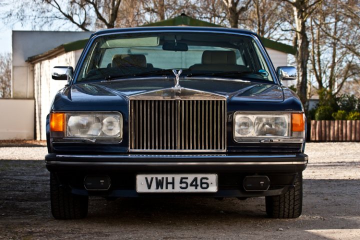 Everything But The Car - Page 1 - Goodwood Events - PistonHeads - The image showcases a vintage luxury vehicle, specifically a Rolls-Royce, parked on a bare ground. The car is positioned in the foreground, with its regal grille and the prominent "VWH" logo immediately drawing attention. The body of the car is painted in a deep blue hue, gleaming under the light. To the left of the image, part of a white shed is glimpsed, suggesting a rural or semi-rural setting. The overall scene is one of elegance and sophistication, harking back to a bygone era of automotive history.