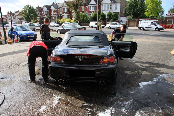 My S2000 review - Page 5 - Honda - PistonHeads - The image depicts a rainy day with two individuals cleaning the rain-soaked rear window of a small, black sports car. The individuals are using what appears to be a squeegee and possibly a cloth on the car's trunk. The car is parked on a street lined with parked cars and buildings in the background. The overall scene suggests that these two people are diligently attending to a task, perhaps protecting the car's window and paint from water damage. The overcast weather adds to the ambiance of the scene, highlighting the downward angle of the raindrops against the reflection on the wet car surface.