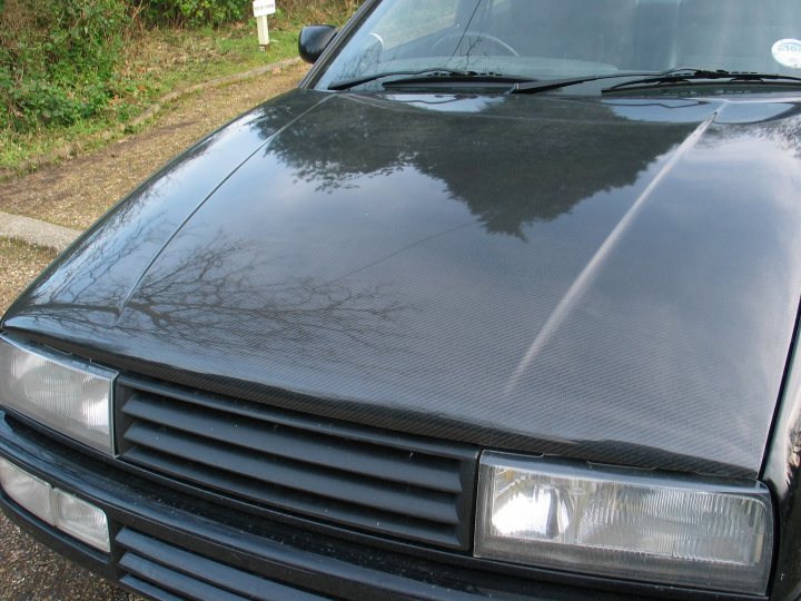 Tweaked Corrado Slightly Pistonheads - The image features the front of a car, with a distinct focus on the hood. The hood displays a black textured surface, possibly a carbon fiber protective wrap. The hood is slightly wrinkled in places, indicating it might have been matte black vinyl or another material applied to its surface. There are no visible vents on the hood, but the father suggests the wrinkles may serve a similar purpose, allowing for heat dissipation. In the background, the car is parked on a gravel driveway amidst some shrubbery, which hints at the possibility of a residential setting.