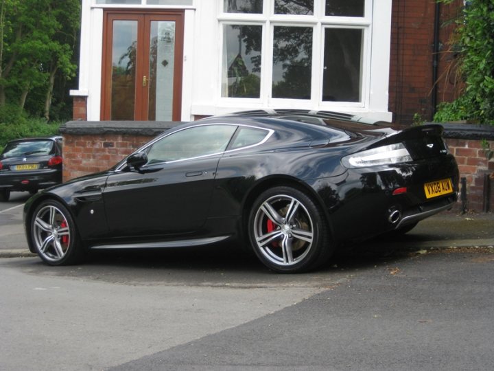 Pistonheads - This image features a sleek, black Aston Martin DB7 convertible parked in front of a two-story house with white framed windows. The car is parked alongside a paved driveway next to a brick wall. Its license plate reads "WW10 FZ". The DB7 is known for its distinctive design, large alloy wheels, and refined driving experience. The scene evokes a sense of elegance and luxury, as befits an Austin Martin.