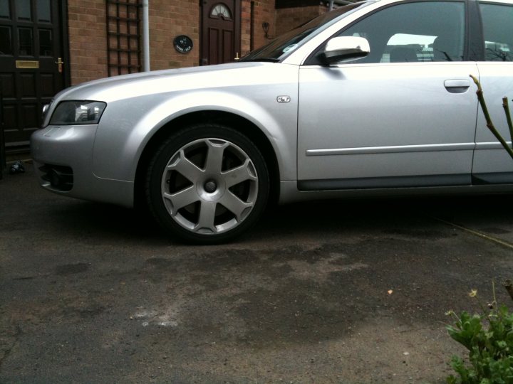 Pistonheads Rscom Managed - This image features a silver Audi car parked on a gray driveway. The car appears to be a four-door model, and it's positioned in a way that shows its profile. A noteworthy detail is a black and silver tire cap with four studs on the front wheel. The car has a modern, sleek design, with achrome grille and headlamps. The driver's side door is open, revealing the interior, which adds a lived-in aspect to the scene. On the right side of the image, there's a small, green plant, partially visible. Overall, the image captures a quiet, residential setting.