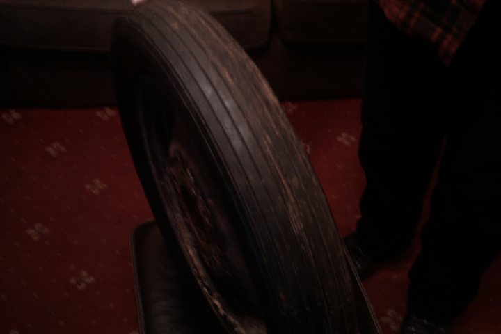 Odd Tyre Sized Pistonheads - The image shows a close-up of an old, worn black rubber car tire. The tire appears to be heavily treaded and is covered with dust and dirt, suggesting it has not been used for a long time. The treads are noticeable with their varying depth and patterns, while the side of the tire appears cracked and aged. The snow pattern in the backdrop contrasts with the indoor setting where the tire is shown, implying a residential or indoor entrance.