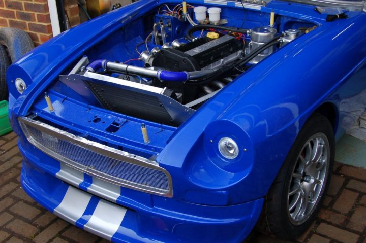 Pistonheads Mgb Special - This image showcases a blue sports car parked in a driveway. The car is characterized by a sleek design with a visible blue and white stripe running down the hood. The dealership bumper and plate are also visible, suggesting that the car is newly purchased or on display. The engine bay of the car is open, revealing the intricate mechanical details, including various components and hoses. The car is equipped with several tires, one of which is prominently placed in the background.