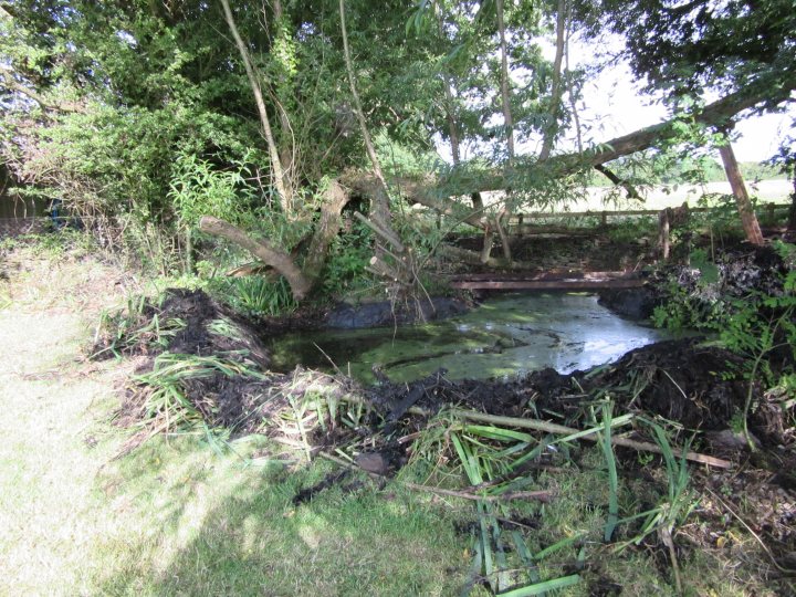 A river with a boat in the middle of it - Pistonheads - The image shows a messy, overgrown outdoor area. There is a deep hole filled with water surrounded by various types of plants and debris. The soil surrounding the hole is eroded and muddy, indicating frequent exposure to rain or other weather elements. From this vantage point, part of a fence is partially visible on the right side of the image. The environment appears to be a neglected corner of a yard or garden, potentially after a severe weather event.