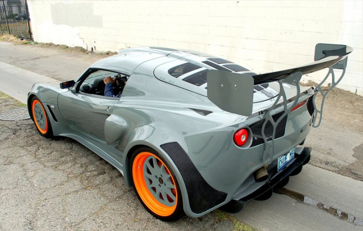 This is a Lotus.. - Page 2 - Elise/Exige/Europa/340R - PistonHeads - The image shows a close-up view of a silver race car positioned on a street alongside a concrete wall. The car's rear wing is prominently displayed, and its wheels have vibrant orange accents. The next to the car is a large dumpster, and there are some plants growing on the concrete surface.