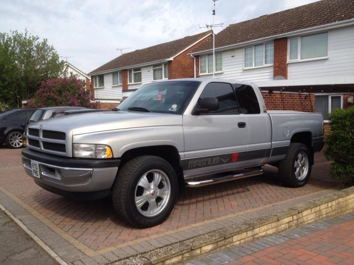 My Dodge Ram. - Page 1 - Yank Motors - PistonHeads - The image depicts a large, four-door, silver Dodge Ram pickup parked on a brick driveway. The truck is parked in front of a house with a white facade and brick details. The vehicle is raised slightly in the front, likely due to its four-wheel drive system, and it features large, chrome wheels. The sky is partly cloudy, suggesting a pleasant day. The image appears to be a photograph taken during the day.