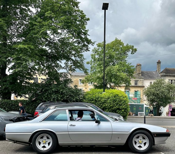 Satan's barge - 1983 Ferrari 400i - Page 8 - Readers' Cars - PistonHeads UK - The image shows a gray sedan parked on the side of a street. Behind the car, there are two people standing near a building with a visible chimney. The sky above is cloudy. The vehicle appears to be an older model, possibly from the 1980s or earlier, and it has a distinctive long hood design characteristic of certain classic cars. There's no text present in the image.