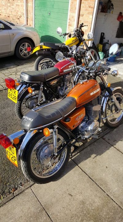 A picture a day....biker banter (Vol 4) - Page 505 - Biker Banter - PistonHeads - The image shows a neatly parked row of three motorcycles. The motorcycles, which have distinct colors and styles, are arranged in a straight line along a concrete driveway. The first bike is gray, followed by a red one, and finally an orange one. The green backdrop suggests the scene might be from a garage or a residential area. There's a car parked on the left side of the image, partially visible, which provides a sense of the scale of the motorcycles.