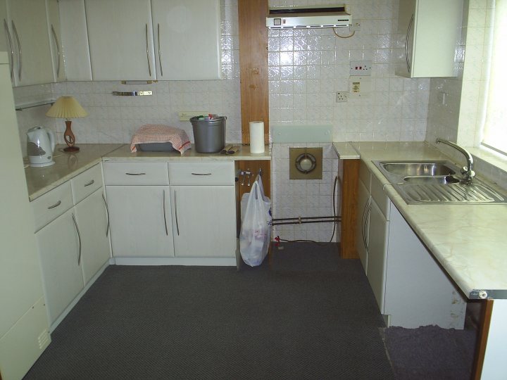 Our house renovation - lovely 1970's style decor - Page 1 - Homes, Gardens and DIY - PistonHeads - The image showcases a neat and tidy kitchen. It features a white cabinette with a vintage design and white butcher block counters. Stark contrast is created between the shades of grey and tile green visible in the wall tiles. A window is situated on the left side, allowing natural light to illuminate the space. On the right side, there is a white sink installed under a window, and a white stove can be seen below the window. The kitchen seems ready for use, with a towel casually placed on the cabinette and a bucket and tools seemingly used for kitchen renovations.