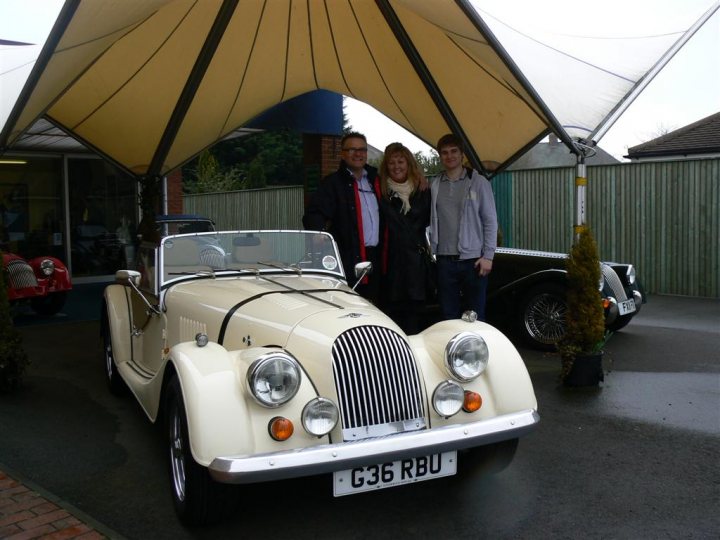 My new 4/4 lowline - Page 1 - Morgan - PistonHeads - The image captures a delightful moment where three individuals stand proudly next to a classic Bentley car under a tent cover. The Bentley is cream-colored and exudes an air of elegance and luxury. The people are smiling as if they are posing for a picture, and they seem to be standing near a building with a green fence. The backdrop is filled with other cars, hinting at a possible car show or exhibition.