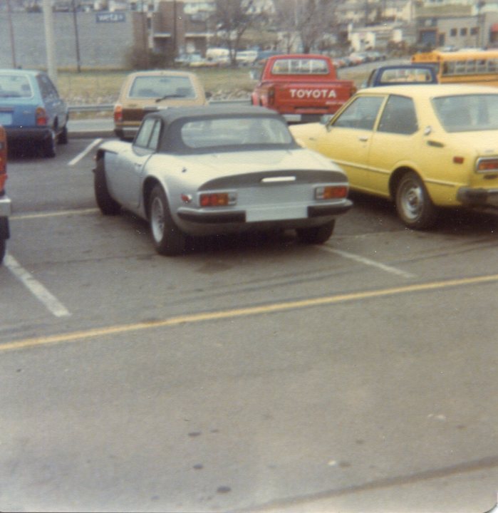 Last 3000S exports to US? - Page 1 - Classics - PistonHeads - The image is a color photograph showing a parking lot. In the center, there's an older, sleek-looking car with a silver body and a black hard top. Surrounding it are various other vehicles, including a bright red minivan, a gold sedan, and a yellow one. The ground is asphalt, lined with parking spaces, and there's a street in the background with additional cars parked along the curb. A prominent "Toyota" emblem is visible on a vehicle behind a white car. The overall scene conveys a casual setting, possibly taken in an urban area.