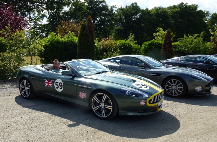 Aston Cars stickered up for LeMans. - Page 1 - Aston Martin - PistonHeads - The image presents a dynamic scene of two cars parked side by side in a forested area. The primary subject of the image is a green luxury convertible, emblazoned with a number "59" and the Aston Martin logo, suggesting it may be a part of a racing team or event. The convertible's top is down, revealing the interior and driver's seat. 

In the driver's seat of the Aston Martin, a man is visible, seemingly comfortable and in control of the car. The interior of the car appears to be well-maintained, with the driver enjoying a clear view of the dashboard, which is illuminated, possibly indicating that the car is running or the lights are on.

The background of the image reveals a serene forest setting, with trees lining both sides of a gravel road that the cars are parked on. This setting provides a natural backdrop to the cars, contrasting with their sleek and sophisticated design.

In the background, there is another luxury car, though its specific make and model are not easily distinguishable from the distance. Its presence suggests that the location might be a gathering spot for enthusiasts of luxury vehicles, or perhaps a