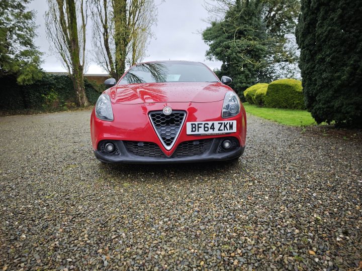 Alfa Romeo Giulietta Quadrifoglio Verde (Launch edition)  - Page 4 - Readers' Cars - PistonHeads UK - The image showcases a red Alfa Romeo Giulietta car parked on a gravel driveway. The vehicle is sleek and has black rims, giving it a sporty look. It's positioned in the middle of the frame with trees visible in the background, suggesting a suburban or rural setting. There are no people or other objects in immediate proximity to the car. The focus of the image is clearly on the car itself, highlighting its design and color.