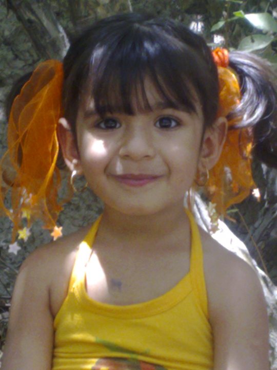 The image features a young girl with a bright smile. She has dark hair adorned with yellow hair accessories. Her dark eyes are the focal point of her face. She is wearing a yellow sleeveless top. The background is blurred, but it appears to be an outdoor setting with some greenery.