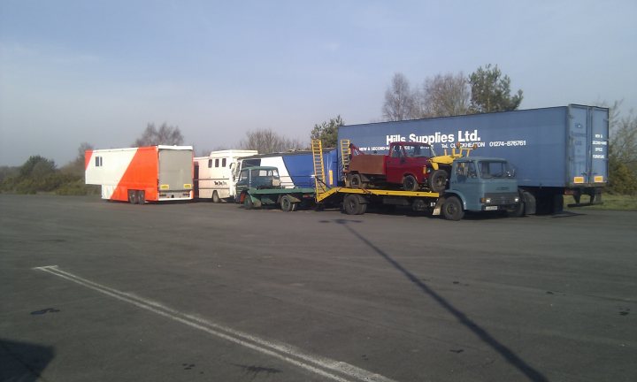 Blackbushe airport turns into a film set for 5 months! - Page 1 - Thames Valley & Surrey - PistonHeads - The image shows a large open space, possibly a parking area, with several delivery trucks parked. The trucks are varied in color, including blue, red, orange, and white, and they are also of different sizes and models. In front of the viewer, there is a red truck with a load bed, and behind it, there is a blue flatbed truck. The ground beneath the trucks appears to be a tarmac surface, and the sky can be seen in the background, suggesting an outdoor location. The trucks are well-maintained and seem ready for dispatch.