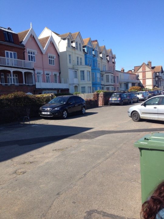 seaside day out, 2 hours from south east essex????? - Page 2 - Holidays & Travel - PistonHeads - This image shows a quiet, residential street with a unique combination of discoveries. Notably, the street is flanked by apartment buildings painted in shades of pink and white, interspersed with blue homes. There are a few cars parked along the street, and a green trash can is visible on the sidewalk. The sky above is clear, suggesting a beautiful day.