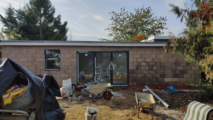 Without planning permission, erection of an outbuilding? - Page 2 - Homes, Gardens and DIY - PistonHeads - The image depicts a construction site with a partially completed house. A building in the background is covered with scaffolding, and there are materials scattered around the area. On the right side of the photo, there's a pile of rubble near a bench, suggesting ongoing work. A blue tarp can be seen on top of one of the structures, possibly for protection against weather elements or to block off an area under construction.