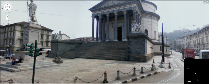 Famous Places Streetview Google Pistonheads - The image depicts a city street scene with a prominent, large, circular, marble building featuring seated statues in the center on a series of steps that ascend to a central pedestal. The structure has classical architectural characteristics such as columns, a dome, and a grand staircase. In the foreground of the image, there is a statue of a soldier and some pedestrian traffic lights. The street itself is flanked by parked cars, and the weather appears to be foggy or overcast, with a pale, grey sky. A few pedestrians and a bus are visible on the right side of the street.