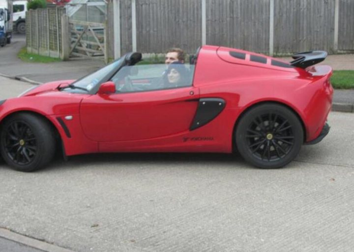 How often do you take the roof off your exige? - Page 1 - Elise/Exige/Europa/340R - PistonHeads - This image features a vibrant, red sportscar driving down a quiet street with trees on one side. The car is sleek and designed with a convertible top, which is down, revealing a clearer view of the interior. In the driver's seat, there are two individuals; they seem to be in a friendly conversation, possibly enjoying a leisurely drive or just arrived at their destination. The street appears to be residential, and there's another vehicle visible in the distance.