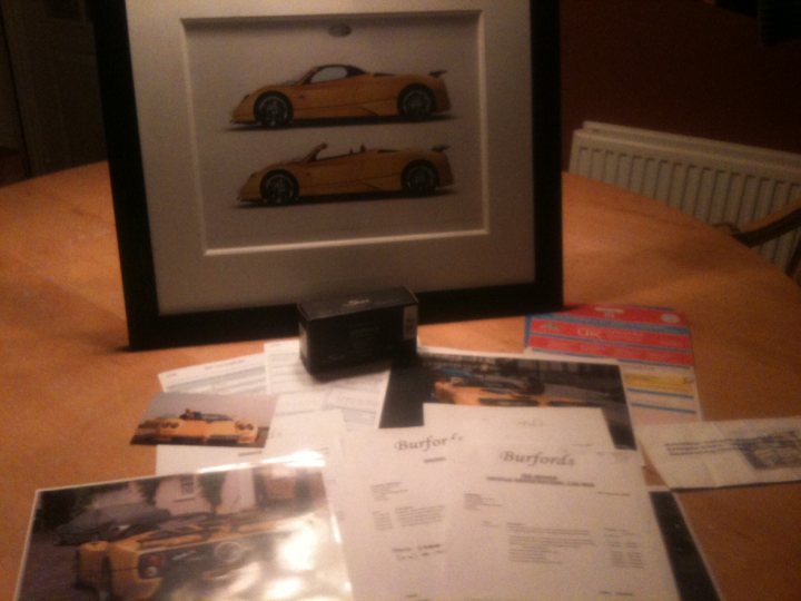 Just bought a Zonda! - Page 6 - Supercar General - PistonHeads - The image depicts a framed picture leaning against a stack of papers and a box on a wooden table. The framed picture shows two cars, possibly luxury models, positioned as if they are racing. Next to the picture, there are several sheets of paper that appear to be related to the framed picture, potentially being the models or makes of the cars. On top of the papers, there is a small black box. In the background slightly blurred, there is a bookshelf with books. The scene suggests a domestic setting, possibly a living room or a study.