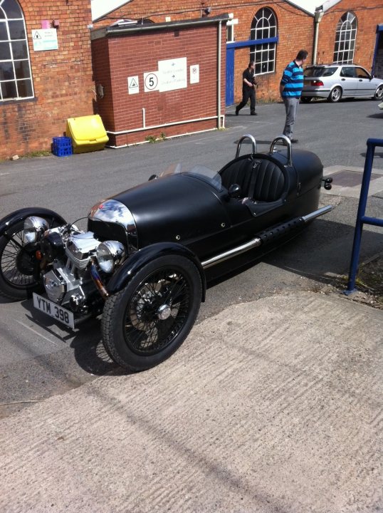 3 wheeler - Page 1 - Morgan - PistonHeads - This image depicts a vintage-style car parked on a street. The car has a classic design, featuring a prominent snub nose, wide-fendered body, and a leather interior visible through the rear seat window. There is a small room with large dormer windows in the background, topped by a metal roof that partially obscures the view of the windows. Pedestrians are visible in the background, and there is a small blue car parked further back on the road. The street appears to be in an urban or suburban area.