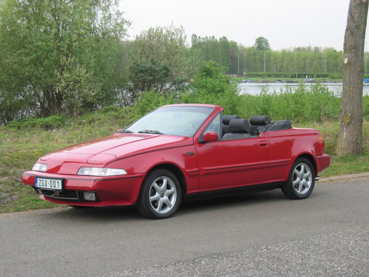 Wedgies Pistonheads Wonderful - The image depicts a vibrant red convertible car parked on the side of a road. The car, a 1995 BMW Z01, is adorned with a German style license plate. It's positioned next to a grass-covered area, possibly a park or a sports field. Behind it, a serene lake or pond is visible, adding to the peaceful ambiance. The sky overhead is overcast, suggesting cooler weather conditions. The overall scene suggests a quiet day in a rural or suburban area.