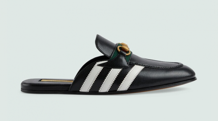 Pistonheads - The image features a black sandal with white stripes on the straps. The sandal is adorned with gold hardware and a green and yellow label, which is part of a larger brand logo. A single brown shoe hangs in the background, suggesting a retail setting. The sandals are positioned centrally against a neutral grey backdrop, emphasizing their design and color scheme.