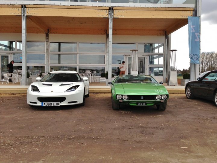 Some nice snaps - Page 1 - Evora - PistonHeads - The image shows a gravel parking surface with three cars placed in front of a covered area which appears to be a pavilion or a covered parking structure. The cars are varied in color: white, green, and black. The white car is equipped with more aerodynamic body modifications, suggesting it might be a high-performance model. The green car is a classic representation of the vintage appearance of a Porsche from the mid-20th century. The black car seems to be a modern, probably a sports or luxury vehicle. The cover provides shelter from the elements and is supported by sturdy metal posts. The building in the background has a modern design, with large glass windows suggesting an open, airy interior that serves as a display or exhibition area. The sky is overcast, with gray clouds obscuring the sky, which gives the scene a neutral background without the distraction of trees or other environmental features. The area is is empty of people, except for one individual standing by the green car. The person is smaller in size due to the perspective, indicating that the image was taken from a distance.