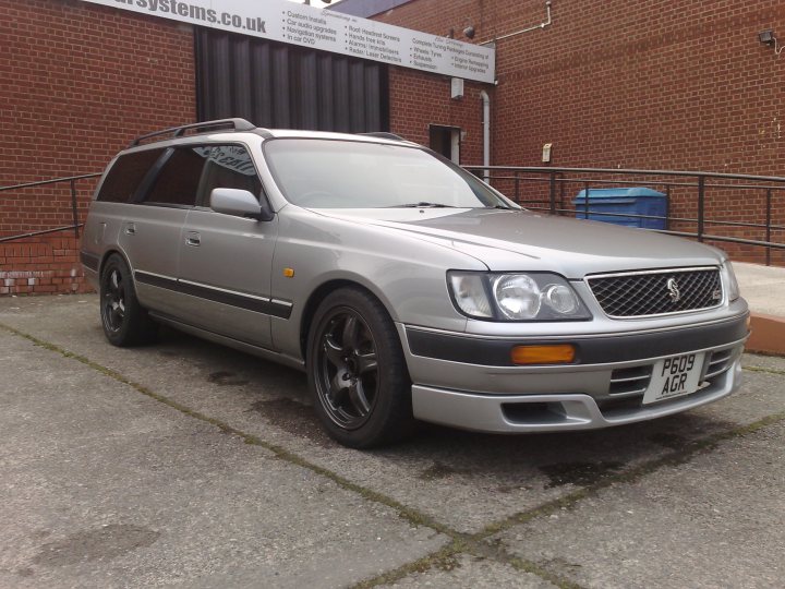 The new Project begins !  - Page 1 - Readers' Cars - PistonHeads - This is an image of a silver car parked near a building. The car features black alloy wheels and a visible emblem on the grill, which along with the license plate suggests that it could be a different brand of vehicle. The building has a sign that reads "??122" on the side, indicating some form of age-restricted access. There is a railing to the right of the car, and further down, there's a blue outdoor bin. The overall setting appears to be an urban environment with a sidewalk and pavement.