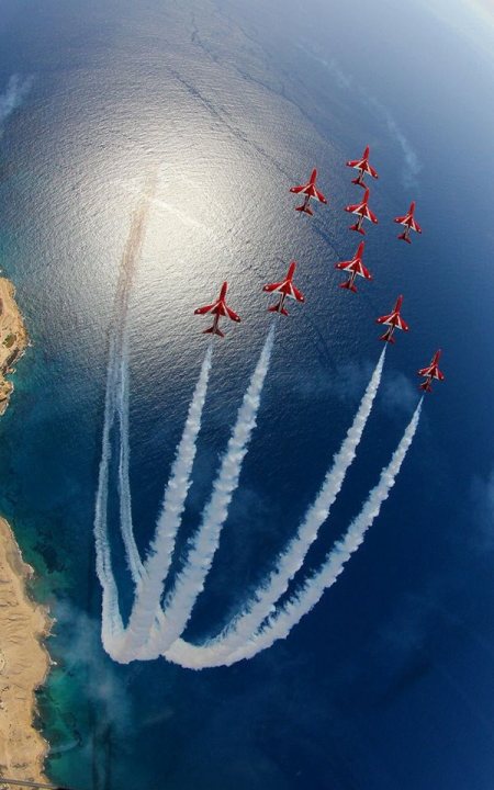 Post amazingly cool pictures of aircraft (Volume 2) - Page 203 - Boats, Planes & Trains - PistonHeads - The image captures a breathtaking aerial view of nine jets flying in a formation over a vast body of water. The planes are painted in vibrant shades of red and white, and they are airborne against a backdrop of a clear sky. The symmetry and precision of their formation suggest a demonstration or an exhibition of aerial acrobatics. The perspective of the photograph adds a sense of depth, emphasizing the scale and magnificence of the scene.