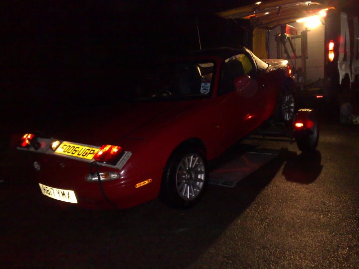 JFReturns Mark 1 MX5 Project - Page 1 - Mazda MX5/Eunos/Miata - PistonHeads - The image shows a vintage red sports car being towed by a white utility truck. The car is predominantly red with the most striking elements being its tail lights that are distinctly separate from the main body of the car. The license plate can be seen hanging from the back of the car. The truck pulling the car has a crowned emblem, but the details are not entirely clear. The tow is taking place at night, indicated by the darkness of the surroundings.