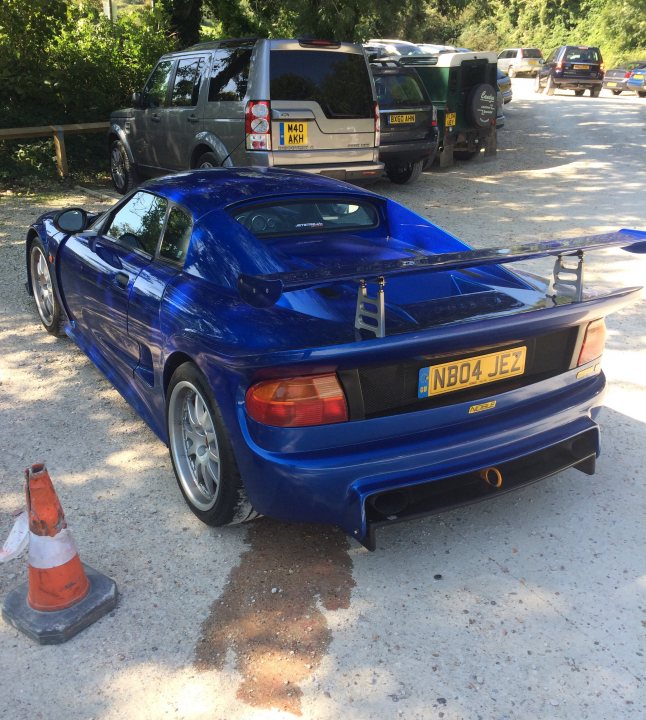 Thar she blows - Page 1 - Noble - PistonHeads - In the image, a vibrant blue racing car is parked on a gravel surface. The car is positioned in the center of the frame, with its rear end facing the camera. The license plate of the vehicle reads "NB04 JEZ". To the left of the car, there's an orange and white traffic cone placed on the ground. In the background, the scene opens up to a parking lot where various cars and trucks are parked, though they are not the main focus of this image. The overall setting suggests an outdoor event or gathering, possibly related to motor vehicles or racing.