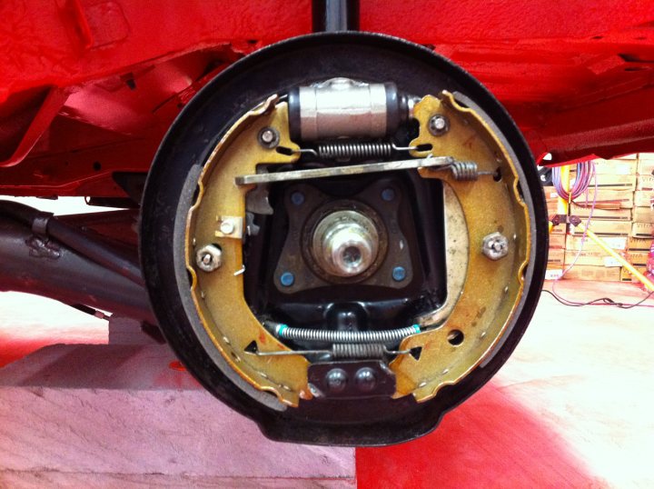 Pistonheads - The image depicts the underside of a red vehicle focusing on the right side of a wheel well. The rim of the wheel is visible, painted in a beige or slightly yellowed color. Inside the wheel well, there is an assembly of metal components with blue and silver bolts. There's also a visible screw that has come loose, potentially leading to damage or malfunction of the vehicle's wheel or suspension system. The wheel itself is partly hidden, and the vehicle appears to be stationary on what could be either a yellow or brown floor.