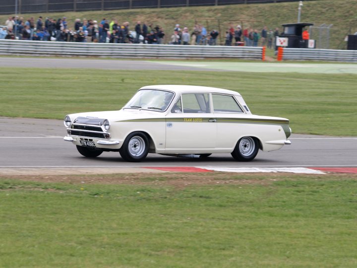 Pistonheads Snetterton - The image features a vintage white car in the foreground on a open road or racetrap. It appears to be a racing model, given its posture and presence at what could be a track. The car's design suggests it is from an earlier era, possibly the early to mid 20th century. In the background, there are several people scattered across the area, some of whom could be spectators or crew members related to the car. The grass and open space suggest a rural or semi-rural landscape, and there seems to be a barrier or fence perhaps demarcating the track boundaries.