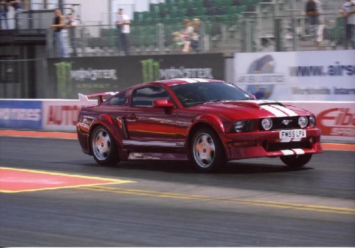 Pistonheads - In the image, a distinctive red car sports a black roof, driving on a raceway. The vehicle has a unique design, characterized by white stripes on its nose and a black rear wing. The raceway itself is set against a backdrop of spectator seating, indicating that a race or event is taking place or about to start. The pace car, similar to the red car, is leading the way down the track.