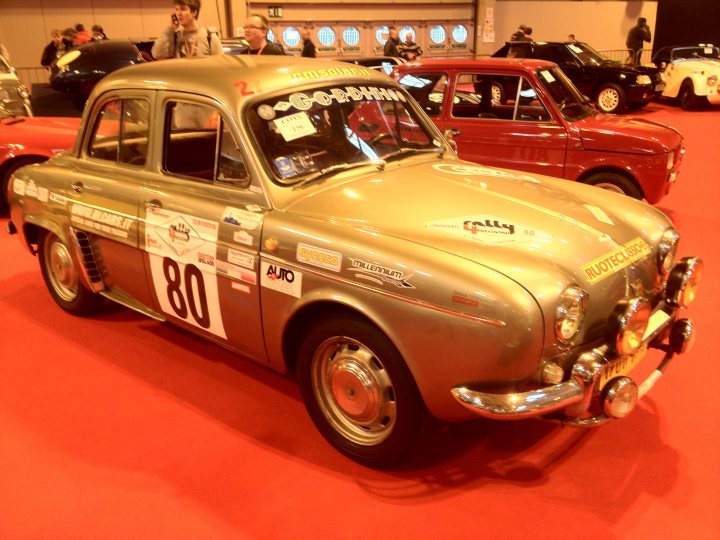 Renault Gordini Dauphine 610 1960 Newbie - Page 1 - French Bred - PistonHeads - The image shows a vintage car standing out on a red floor, possibly at a car show. The car has a metallic golden-yellow finish and features prominent headlamps, the text "80" painted on the side, and a log on the hood. Its old-style design suggests it is a classic model. In the background, another vintage car can be seen, indicating that more antique vehicles are likely to surround this one at this event. There are people in the background, likely admirers or staff at the event, but they are not the focus of the image.