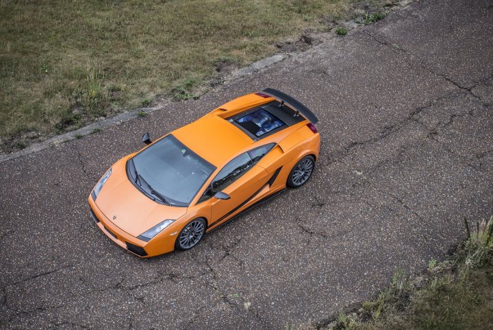 Superleggera - Page 1 - Gallardo/Huracan - PistonHeads - The image depicts a vibrant orange sports car positioned in the center of a split road. The car is in motion on a rough asphalt surface, with the driver's side visible. The vehicle features a distinct aerodynamic design and large black wheels that contrast with its bright color. Adjacent to the road, there is a grassy field that complements the natural surroundings, setting the stage for a dynamic and lively drive.