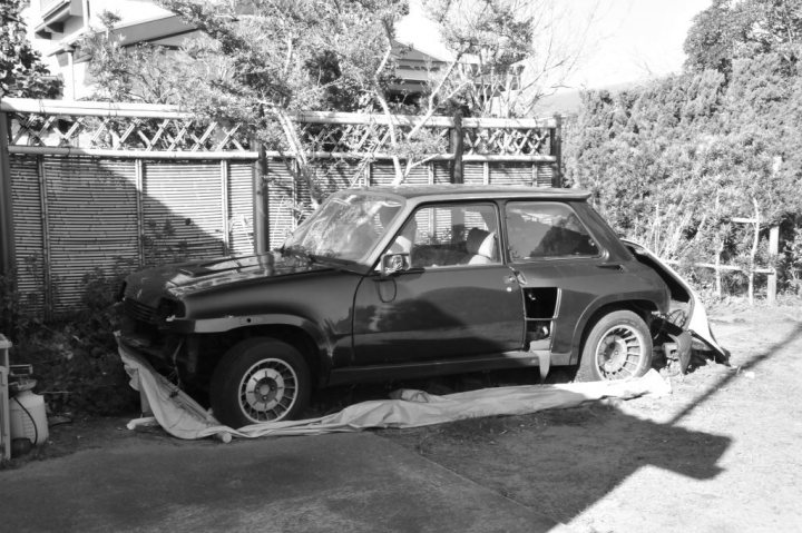 Classics left to die/rotting pics - Page 481 - Classic Cars and Yesterday's Heroes - PistonHeads - The image depicts a vintage car parked next to a fence on a stone surface. It's an older model with a glossy finish, giving it a distinguished appearance. Underneath, it appears it has been involved in an accident, with visible damage to the front-right area. There are no visible texts or distinctive features that provide specific information about the vehicle or its location. The photo is in black and white, which enhances the timeless quality of the scene.