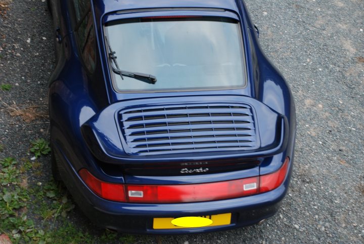 Is this car a wide or narrow body 993? - Page 1 - Porsche General - PistonHeads - The image presents a serene outdoor scene featuring a **blue car** parked on what appears to be a **gravel surface**. The car, a **Porsche 456**, is viewed from behind, emphasizing its distinctive design. The window of the back window is up, and it reflects a **red light**, suggesting that the car may have been parked there recently. In the background, there are some **patchy green plants**, adding a touch of nature to the scene. The overall atmosphere is quiet and peaceful, with the car looking well-maintained and ready for a drive.