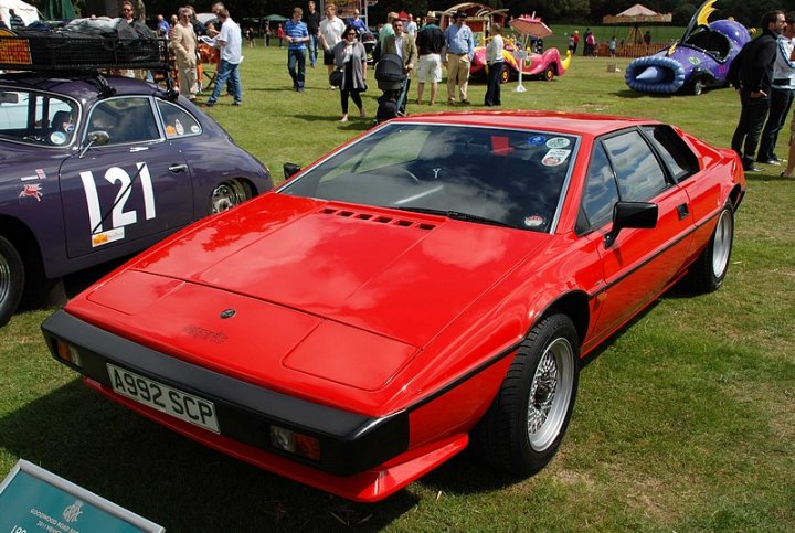 GRRC Lunch Club - Page 1 - Goodwood Events - PistonHeads - The image captures an outdoor scene with a vibrant red DMC DeLorean car parked on grass. This car, famously depicted in the "Back to the Future" movie franchise, is the center of attention. It's positioned between another purple car and a large orange kite, adding a splash of contrasting colors to the scene. Surrounding these vehicles are several onlookers, their presence adding a sense of scale to the image. The setting appears to be a fair or event, as indicated by the numerous people and festival-like atmosphere.