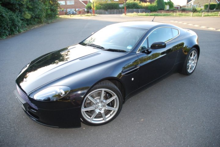 Pistonheads Promised - The image features a black Aston Martin car parked in a sunlit area. The car has a sleek, shiny exterior, and the reflection on its glassy surface highlights its polished design. The car is parked on the outer side of a parking lot, with a partially visible brick building and a street visible in the background. The shadow of the car is cast on the ground, indicating the sun is in the sky.