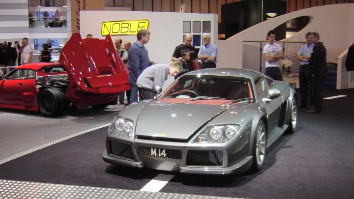 Pistonheads - The image captures a sophisticated setting featuring a silver sports car on display. The car is sleek and appears to have a glossy finish, with its prominent front grille and well-defined headlights that contribute to its stylish design. Surrounding the car, there are several well-dressed individuals who seem to be observing the car attentively or waiting for their turn to explore it. The presence of a red car in the background suggests that other vintage cars may also be part of the exhibition or sales event. The atmosphere appears to be formal and exclusive, with the showcased car and the attendees' attire suggesting a high-end or luxury market.