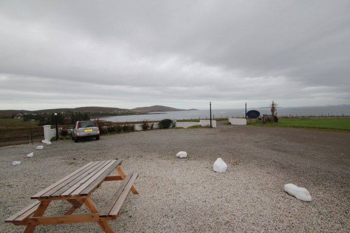 Highlands - Page 69 - Roads - PistonHeads - The image shows a large, empty parking lot with an unoccupied wooden picnic table at the forefront. The ground is made of gravel, and the lot appears to be near a body of water, as suggested by the presence of water alongside the parking area. There are a couple of distant figures that seem to be walking in the far background, surrounding the van, which indicates someone's presence. There's also a piece of wooden outdoor furniture or a similar item that's not fully visible in the scene. The sky is overcast with gloomy, cloudy weather suggesting a cool or possibly rainy day.