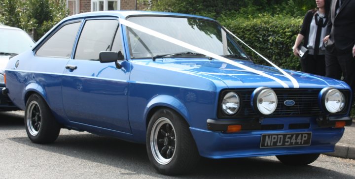 Wedding Ideas Pistonheads - The image showcases a vibrant blue classic car, possibly a Ford, parked on a street. The vehicle features distinctive round headlights and a white vinyl top and stripe that run the length of its body. A white ribbon adorns the front, indicating it might be on display or for a special occasion. In the background, there are other cars parked and a glimpse of a person, suggesting a public setting. The classic nature of the car and the presence of the statement top and stripe suggest it might be a pride and joy for its owner, possibly even at events like car shows.